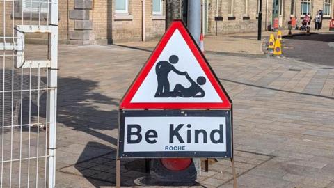 A triangular road sign, with a white face, red edgings and black text, reads 'Be Kind' and shows a monochrome cartoon illustration of a person helping another off the floor.