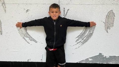 A young boy with brown hair stands with his arms outstretched next to a wall. The wall has marks on it which loosely look like wings. The boy is wearing a black zip up jacket and black shorts. 