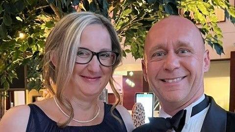 Amie Henson and Dan Lee pictured together. Amie has blonde hear and black glasses and is wearing a dress with a necklace. Dan wears a tuxedo and is bald, he is smiling.