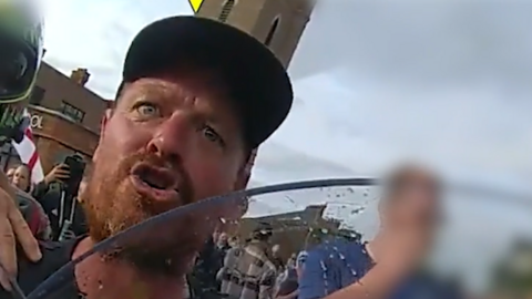 A man wearing a black cap alleged to be Lee Gower. He appears to be shouting at a police officer who is behind a riot shield.