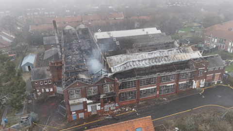 A drone image of a large red-brick factory building, with most of the roof space collapsed after a fire. Smoke rises from the exposed building, which is surrounded by terraced houses and gardens