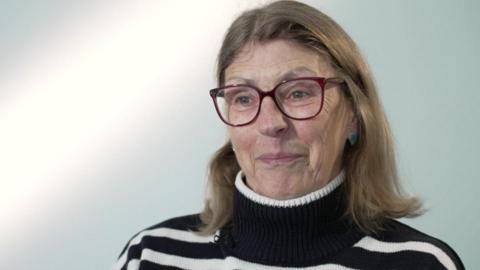 A woman with short light brown hair and glasses smiling to the camera. She is sitting on a chair against a white backdrop and wearing a black and white striped jumper.