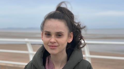 Freya is standing on a coastal path in Rhyl. The beach and sea is behind her. She is looking straight at the camera and has a small smile. She has brown hair that has been tied back because of the windy weather. The sky is very grey and Freya is wearing a large puffer jacket to keep warm. 