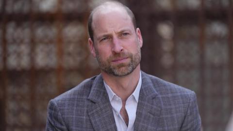 Prince William, wearing a light-coloured striped shirt without a tie and a checked suit jacket looks on with a neutral expression