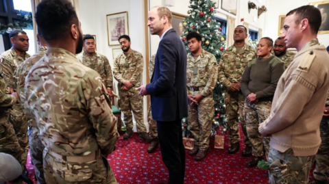 Prince William with a circle of Fijian soldiers in uniform surrounding him in a large room with a Christmas tree in the background