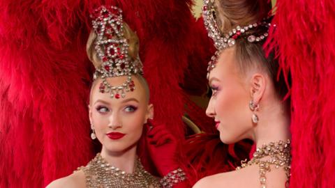 Amelia stood looking in the mirror adorned in red jewels and feathers 