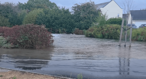 A road is entirely submerged by water. There are bushes around it and houses in the background, beyond the hedgerows.