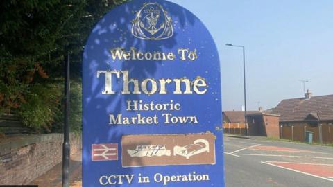 A large blue sign on a suburban street which reads "Thorne Historic Market Town"  then has a rail red and white logo, and a brown heritage logo with a boat and a bed, then underneath the words CCTV in operation.