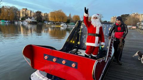 Santa Claus stands aboard a red sleigh mounted on a small boat moored on the River Thames, waving towards the camera, while another person stands on the riverside pontoon beside the vessel.