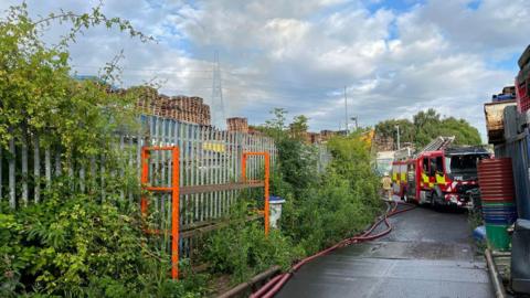 A fire engine parked in front of the waste disposal sites. Behind a metal fence are stacked hundreds of wooden pallets