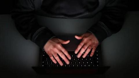 A dark scene looking down on someone dressed in a black long sleeve top leaning with their hands hovering over the keypad of a black laptop.