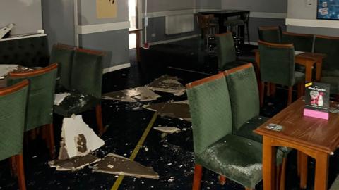 Broken ceiling panels on the floor of a community centre in Yate, with green chairs and tables covered in dust.