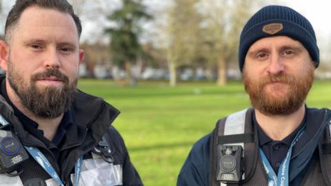 A man with a black beard and another man with a ginger beard both wearing high vis vests and body cameras smile at the camera