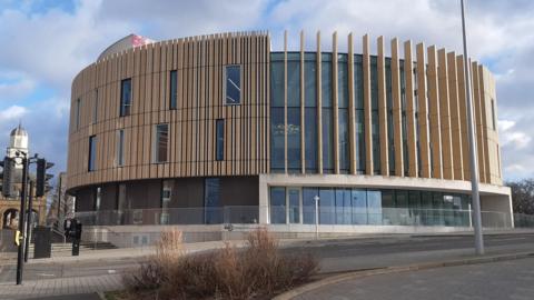 The outside of The Word which is a large, round, modern building with wooden panels lining the windows on the upper floors. There is a large glass entrance on the bottom floor and a glass railing in front of it.
