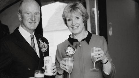 Photo of Tacey and David at a wedding. It is a black and white photo. David is wearing a suit and tie and holding a can or beer and a glass of beer in both hands. Tacey is smiling to the camera holding wine glasses in both hands.