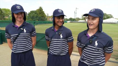 Three boys dressed in navy and white Wimbledon uniforms standing in front of a tennis court.