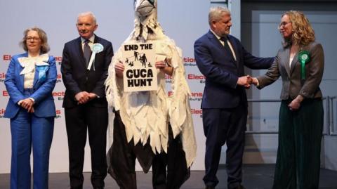 A group of people wearing suits and one dressed as a bird stand on stage