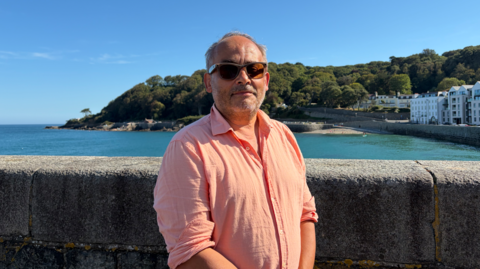 Man wearing glasses and a Light salmon coloured linen shirt, centre of the shot, stood in front of Havalet Bay. It is a sunny day.