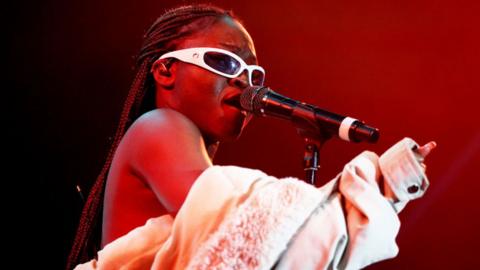 Sampa the Great singing at a microphone. She is wearing white-framed sunglasses and her white jacket is falling off her shoulders.