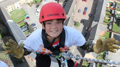 Jacob, a boy who is attached to a harness and rope and is abseiling down the National Lift Tower in Northampton. He is wearing yellow fabric gloves and a red helmet. He is wearing a white T-shirt and is looking at the camera and smiling.