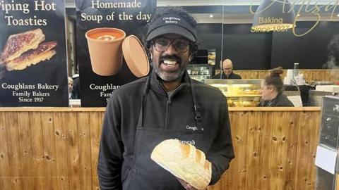 Romesh Ranganathan wearing a black cap and black overall and holding a loaf of bread standing behind the counter at Coughlans Bakery in Dorking