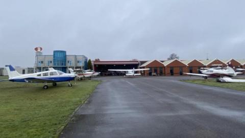 A number of planes parked on grass alongside an area of tarmac with a number of brick buildings in the background.