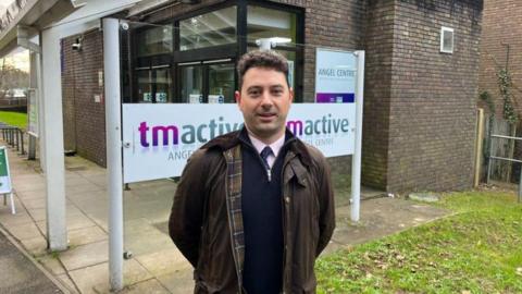 A man stands outside a lesiure centre. It is made of grey bricks. He is wearing a brown Barbour jacket.