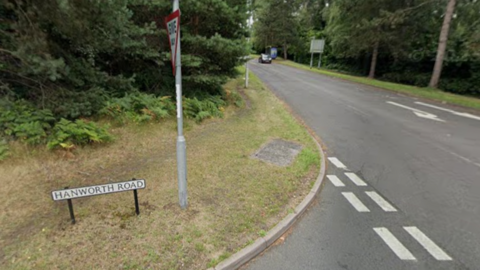 A road sign on a grass verge next to a Give Way sign at a T junction says Hanworth Road. There is no visible pavement on the main road, which is Ringmead.