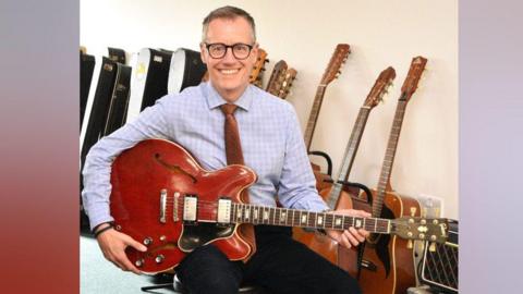 Alexander Clement sits on a chair, he is wearing a blue shirt and brown tie, and has brown hair and black glasses. He is holding a red guitar