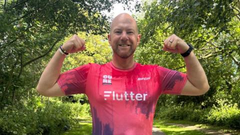 A bald man wearing a pink sports shirt grins at the camera as he flexes his arms. The shirt has the Cure Leukaemia logo on it.