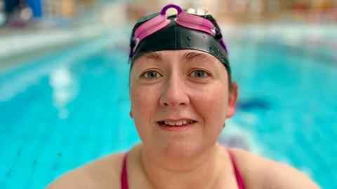 Gemma Stevenson is looking directly at the camera and smiling slightly while wearing a black swimming cap and pink goggles, which are off her eyes and resting on her head. She is holding onto the side of the pool.  Behind her the water is a bright blue and there are no other people in the swimming pool. There are droplets of water on her nose and the edges of her swimming cap. The straps of her swimming costume are pink and can be seen on shoulders, just in frame of the camera. 