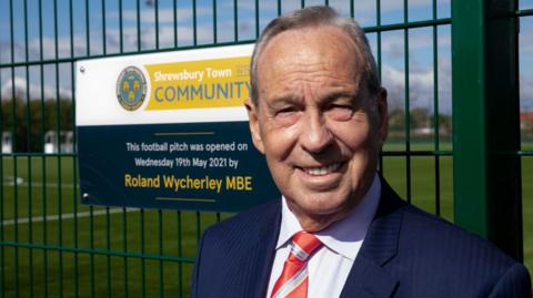 Shrewsbury Town chairman and owner Roland Wycherley stands in front of green metal fence at the club's training ground. He is wearing a navy blue suit with a white shirt and red and white striped tie.