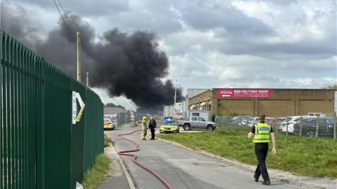 Black smoke is seen coming from behind a Bed Factory Shop building. A police officer walks away from the camera. Two police cars are parked ahead and a firefighters is talking to a man wearing black.