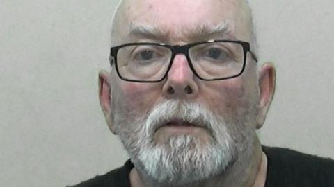 Mugshot of a bald man with a white beard and glasses.