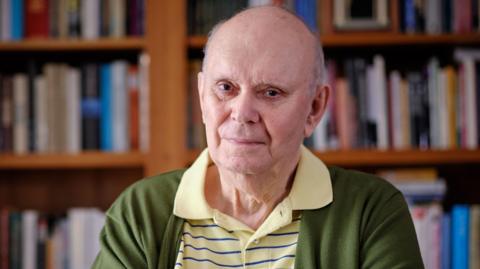 A bald man wearing a light yellow polo shirt with thin navy horizontal stripes, and a green cardigan over the top. He is sat in a library or bookshop, with shelves of books blurred in the background.