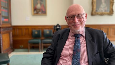 Max Caller, lead commissioner at Birmingham City Council, sits inside an ornate room at the council's HQ. He looks at the camera and is wearing a pink shirt and dark grey suit jacket with a flower patterned tie.