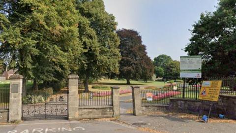 A stone fence with metal railings and a metal gate at the entrance to a park with ornamental flower beds and trees. There is a green and white sign at the top of one of the sections of railings and a yellow sign advising work is being carried out