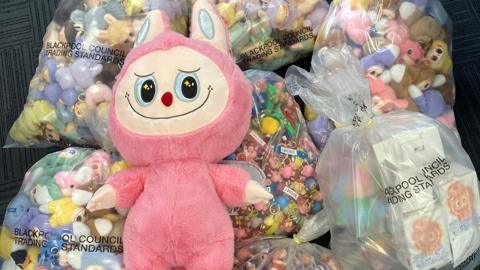 A large fake stuffed Labubu toy resting on top of bags of smaller dolls that have been seized. The large toy is pink with  ears like a rabbit and a pale embroidered face with zigzag teeth, big eyes and a small red nose