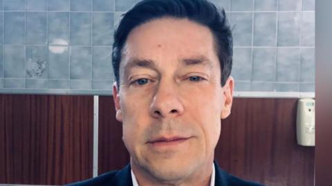 A mirror selfie of a man in a bathroom with blue tiles. He is in his mid-50s and has a half-smile expression. Although the photo is cropped you can tell he has a suit, shirt and tie on and is freshly shaven.