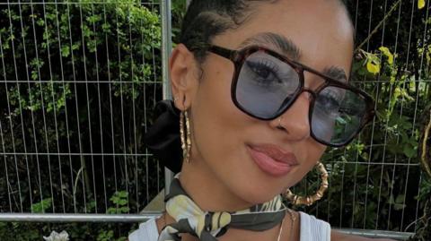 A woman is wearing large sunglasses and earrings, and is looking at the camera.