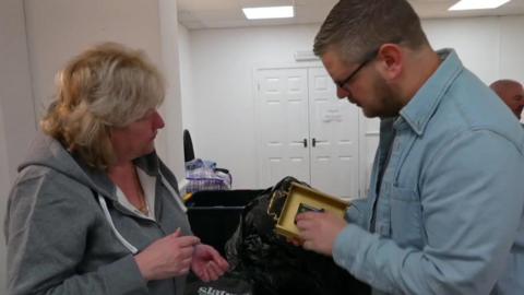 A woman with blonde hair and a grey zip-up hoodie converses with Hames Wood, who has short light brown hair and glasses and is wearing a light blue denim jacket. He is holding a small standalone clock in a bronze case. 