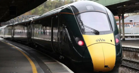 Yellow front of train coming towards the camera with two red lights lit and dark green carriages behind -railway platforms either side.