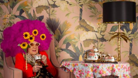 The image shows an elderly woman sat in a chair, wearing a bright pink wig with five sunflowers in them. She is wearing a large pearl necklace and sipping on some tea. The wallpaper in the background is covered in leaves and a black and gold lamp stands on a table to the right, along with an ornate tea pot and cups.