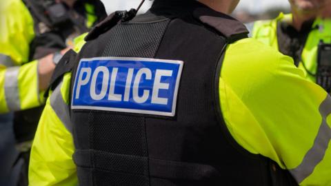 Close up of the back of police officer's black jacket, which has the letters police in white written on. There are two other officers in the distance but you cannot see their faces.