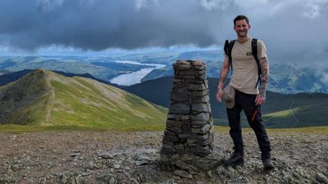 Mr Kendall is pictured on top of a mountain in the Lake District. He stands next to a cairn