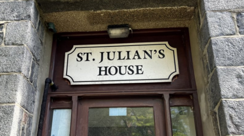 The front door sign for St Julian's House. It is a cream colour above a brown-framed door.