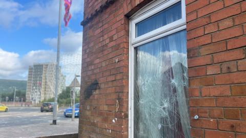 Smashed window on a red brick building, roundabout and tow block in the background 