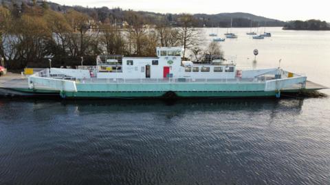 A stock image of the Mallard moored at Ferry Nab in Windermere. The empty ferry has a green hull and white deck. Sailing boats and hills are visible in the background.