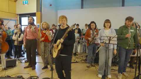 Ed Sheeran singing at a microphone. He is in the foreground wearing all black with a light-coloured guitar in his hands. In the background is a performance group playing instruments.