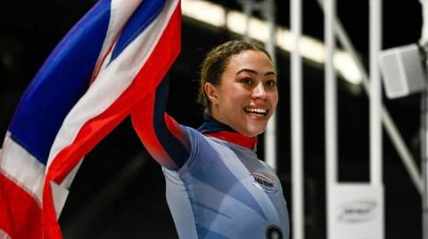 Tabby Stoecker smiling and waving a union jack flag. She has dark hair, tied back, and is wearing a lycra kit adorned with a pattern similar to that on the flag.